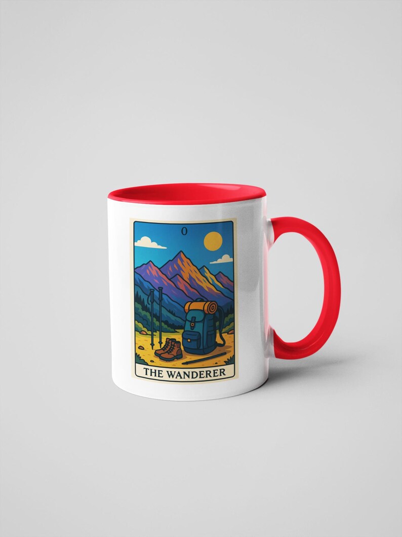 May include: White ceramic mug with a red interior and handle. The mug features a colorful illustration of a mountain scene with hiking gear and the text "THE WANDERER". The design includes a backpack, boots, and hiking poles.