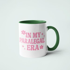 Op de afbeelding: Witte keramische mok met een groene binnenkant en handvat. De mok heeft de roze tekst "IN MY PARALEGAL ERA" met bloemen- en sterrenafbeeldingen. Een leuke en stijlvolle mok voor paralegals.