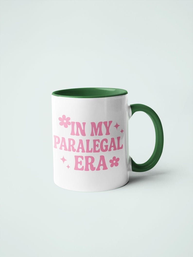 Op de afbeelding: Witte keramische mok met een groene binnenkant en handvat. De mok heeft de roze tekst "IN MY PARALEGAL ERA" met bloemen- en sterrenafbeeldingen. Een leuke en stijlvolle mok voor paralegals.