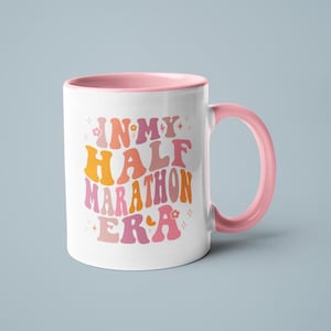 May include: White ceramic mug with a pink interior and handle. The mug features the text "IN MY HALF MARATHON ERA" in retro orange and pink lettering, with floral accents. A fun and motivational item for runners.
