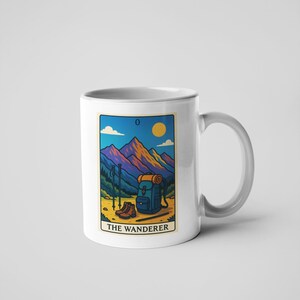 May include: White ceramic mug featuring a colorful illustration of a mountain scene with hiking gear. The design includes a backpack, boots, and trekking poles, with the text "THE WANDERER" at the bottom. The background shows a mountain range, a sun, and a blue sky.