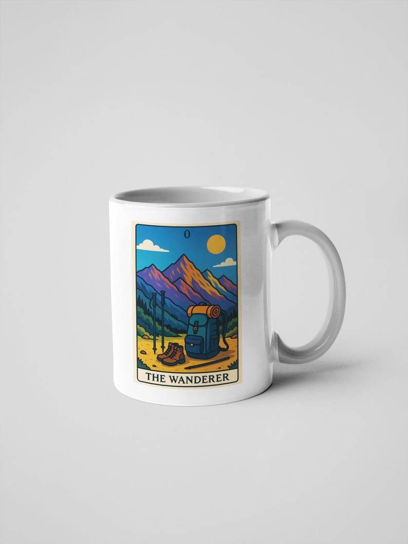 May include: White ceramic mug featuring a colorful illustration of a mountain scene with hiking gear. The design includes a backpack, boots, and trekking poles, with the text "THE WANDERER" at the bottom. The background shows a mountain range, a sun, and a blue sky.
