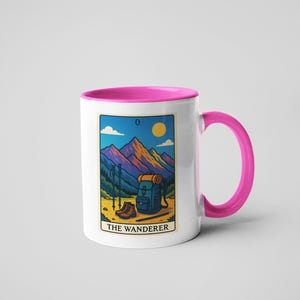May include: White ceramic mug with a pink interior and handle. The mug features a rectangular design with a mountain landscape, hiking boots, backpack, and the text "THE WANDERER". The design is in shades of blue, orange, and yellow.