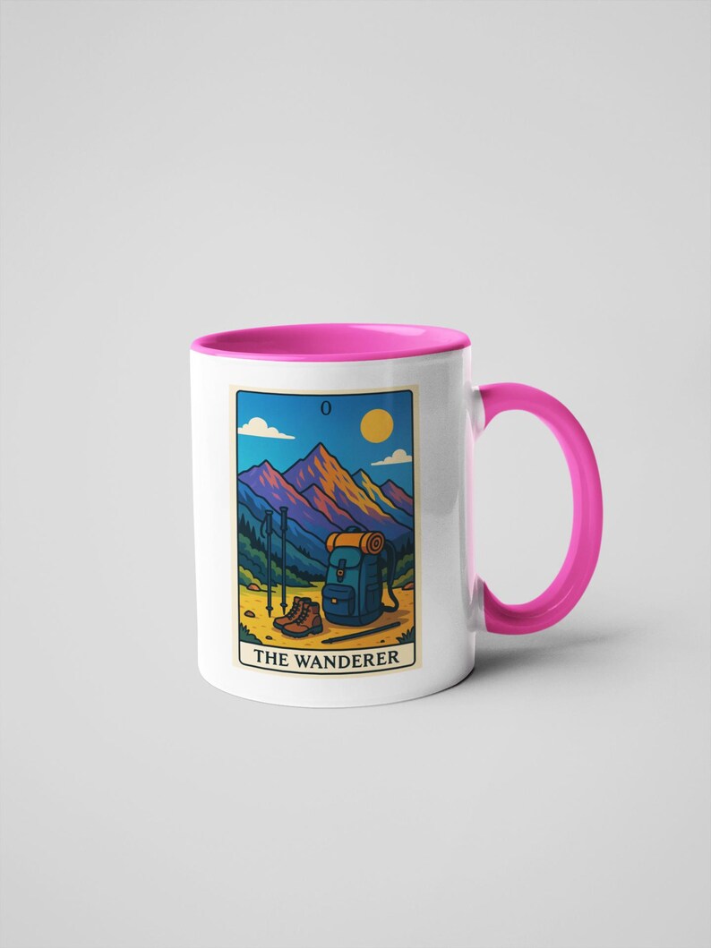 May include: White ceramic mug with a pink interior and handle. The mug features a rectangular design with a mountain landscape, hiking boots, backpack, and the text "THE WANDERER". The design is in shades of blue, orange, and yellow.