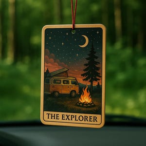 May include: Rectangular air freshener with a camping scene. The image shows a yellow camper van, campfire, and a starry night sky with a crescent moon. The text "THE EXPLORER" is at the bottom. The air freshener hangs from a red string.