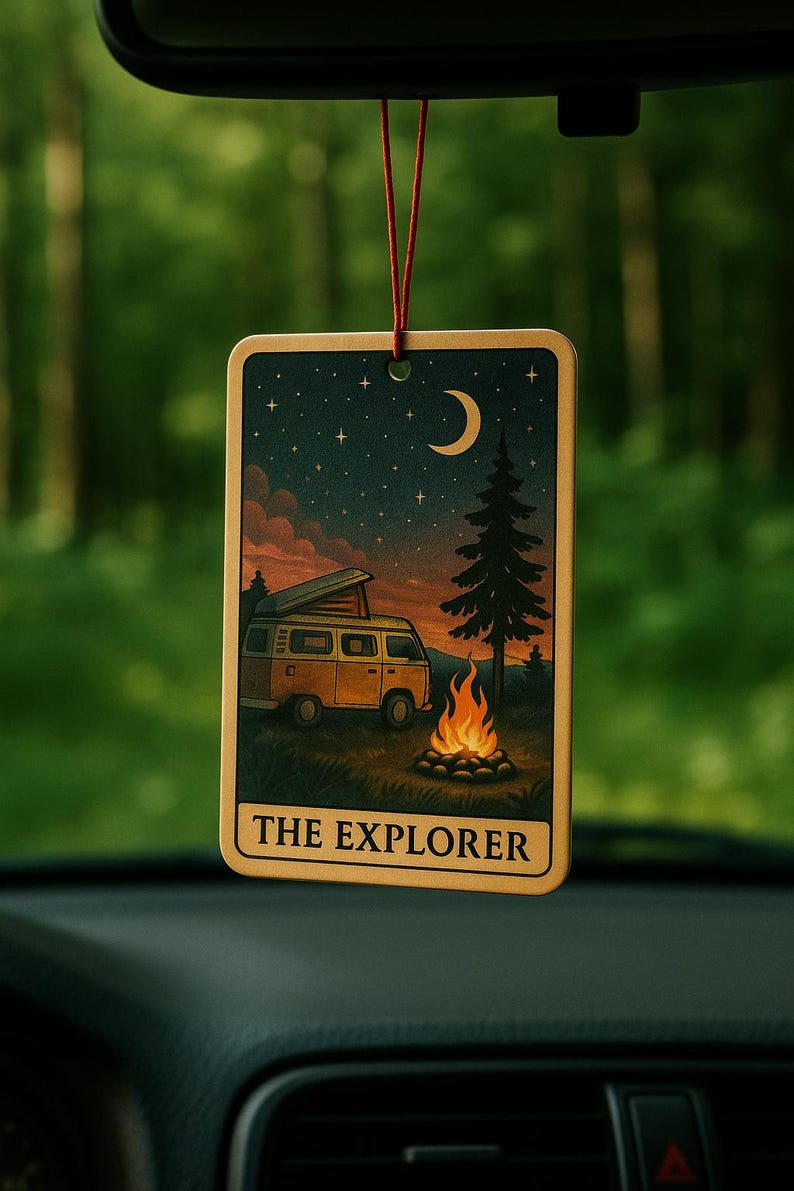 May include: Rectangular air freshener with a camping scene. The image shows a yellow camper van, campfire, and a starry night sky with a crescent moon. The text "THE EXPLORER" is at the bottom. The air freshener hangs from a red string.