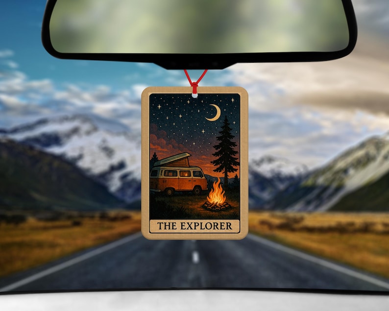 May include: Wooden car air freshener with a scenic illustration of a camper van parked near a campfire under a starry night sky. The text "THE EXPLORER" is at the bottom. The air freshener hangs from a red string.