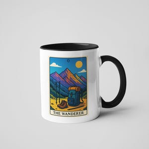 May include: White ceramic mug with a black interior and handle. The mug features a colorful illustration of a mountain scene with hiking gear, a backpack, and the text "THE WANDERER". The design includes a sun, mountains, and hiking poles.