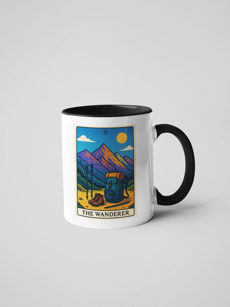 May include: White ceramic mug with a black interior and handle. The mug features a colorful illustration of a mountain scene with hiking gear, a backpack, and the text "THE WANDERER". The design includes a sun, mountains, and hiking poles.