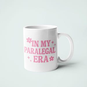 Op de afbeelding: Witte keramische mok met de roze tekst "IN MY PARALEGAL ERA", versierd met bloem- en stermotieven. De mok heeft een gebogen handvat en staat tegen een lichtblauwe achtergrond.