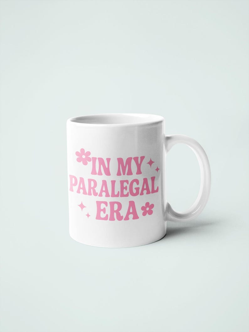 Op de afbeelding: Witte keramische mok met de roze tekst "IN MY PARALEGAL ERA", versierd met bloem- en stermotieven. De mok heeft een gebogen handvat en staat tegen een lichtblauwe achtergrond.