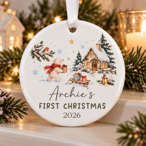 May include: A round white ceramic Christmas ornament featuring a polar bear in a red scarf carrying a gift, a snowy cottage, and a robin. The text reads Archie's FIRST CHRISTMAS 2026. A white ribbon is attached to the top for hanging purposes.