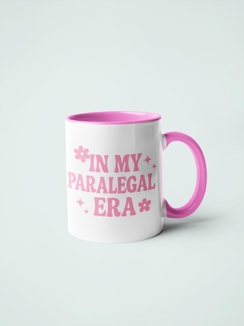 Op de afbeelding: Witte keramische mok met een roze binnenkant en handvat, met de roze tekst "IN MY PARALEGAL ERA". De tekst is versierd met bloemen en sterren.