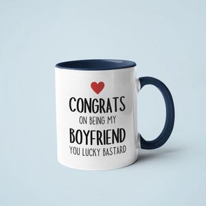 A white mug with a blue handle, displaying the text "CONGRATS ON BEING MY BOYFRIEND YOU LUCKY BASTARD" along with a red heart symbol.