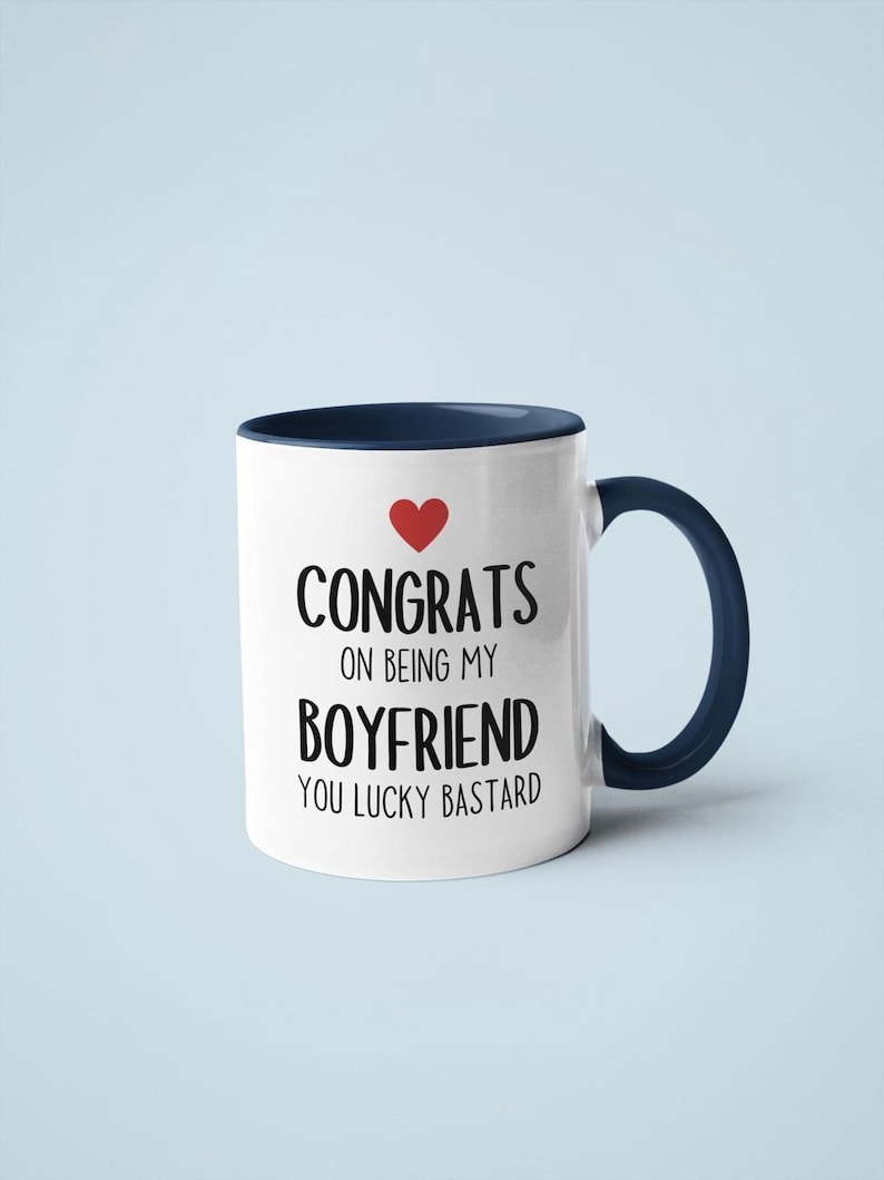 A white mug with a blue handle, displaying the text "CONGRATS ON BEING MY BOYFRIEND YOU LUCKY BASTARD" along with a red heart symbol.