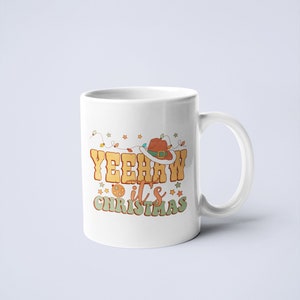 May include: A white ceramic mug featuring a graphic design with the text YEEHAW it's CHRISTMAS in orange and green lettering. The design includes a brown cowboy hat, a string of colorful holiday lights, and small star shapes in orange and green.
