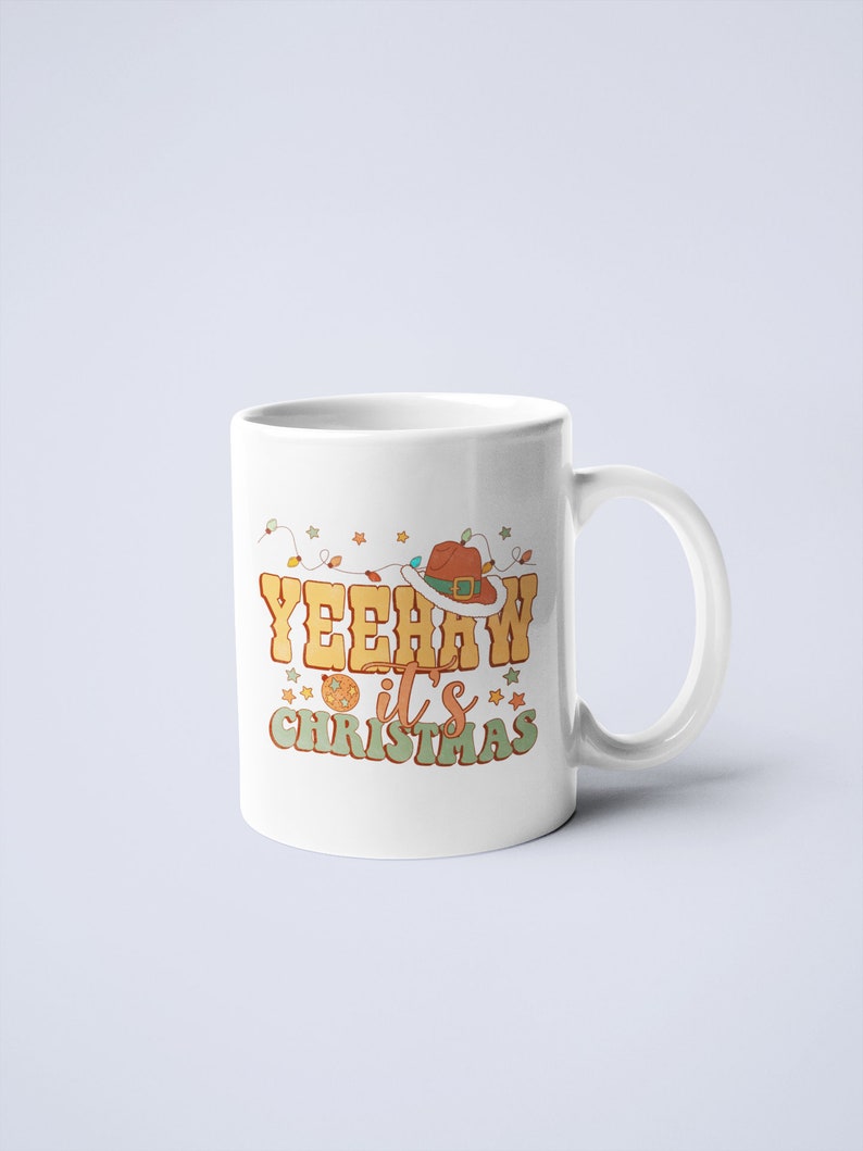 May include: A white ceramic mug featuring a graphic design with the text YEEHAW it's CHRISTMAS in orange and green lettering. The design includes a brown cowboy hat, a string of colorful holiday lights, and small star shapes in orange and green.