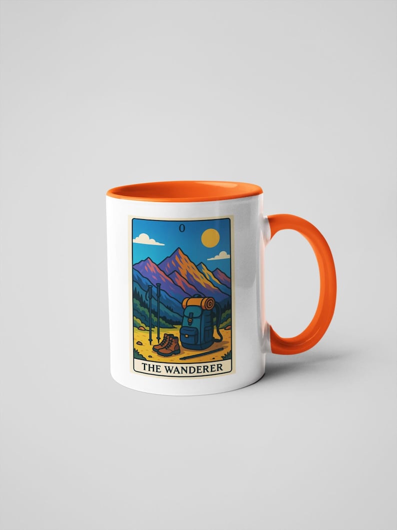 May include: White ceramic mug with an orange interior and handle. The mug features a tarot card design with a mountain landscape, backpack, hiking boots, and the text "THE WANDERER".