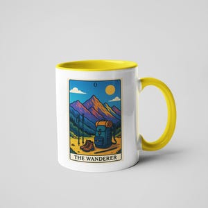 May include: White ceramic mug with a yellow interior and handle. The mug features a colorful illustration of a mountain scene with hiking gear and the text "THE WANDERER". The mug is a standard 11 oz size.