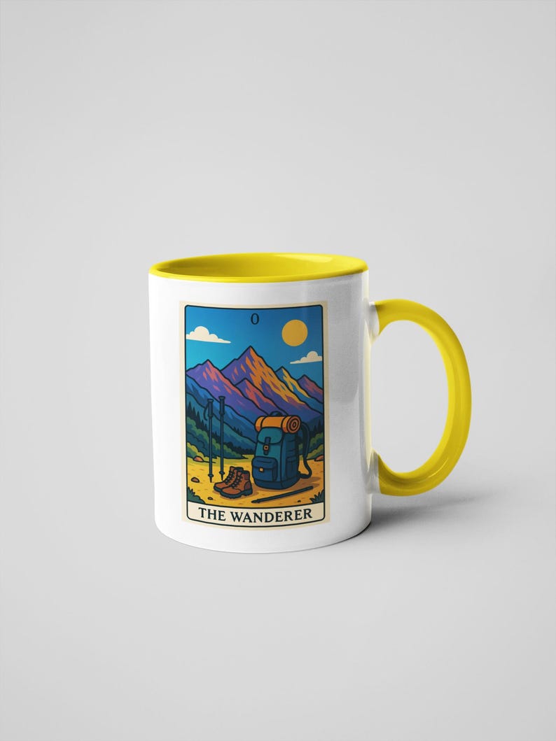 May include: White ceramic mug with a yellow interior and handle. The mug features a colorful illustration of a mountain scene with hiking gear and the text "THE WANDERER". The mug is a standard 11 oz size.