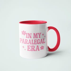 Op de afbeelding: Witte keramische mok met een rode binnenkant en handvat. De mok heeft de tekst "IN MY PARALEGAL ERA" in roze, met bloem- en steraccenten. Een eenvoudig, maar stijlvol ontwerp.