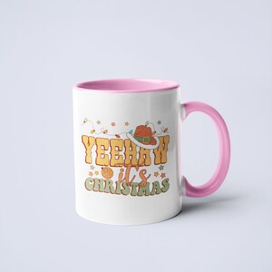 May include: A white ceramic mug with a pink interior and handle. The front features a graphic with the text YEEHAW it's CHRISTMAS in yellow and green letters, accompanied by a brown cowboy hat, a disco ball, and colorful string lights.