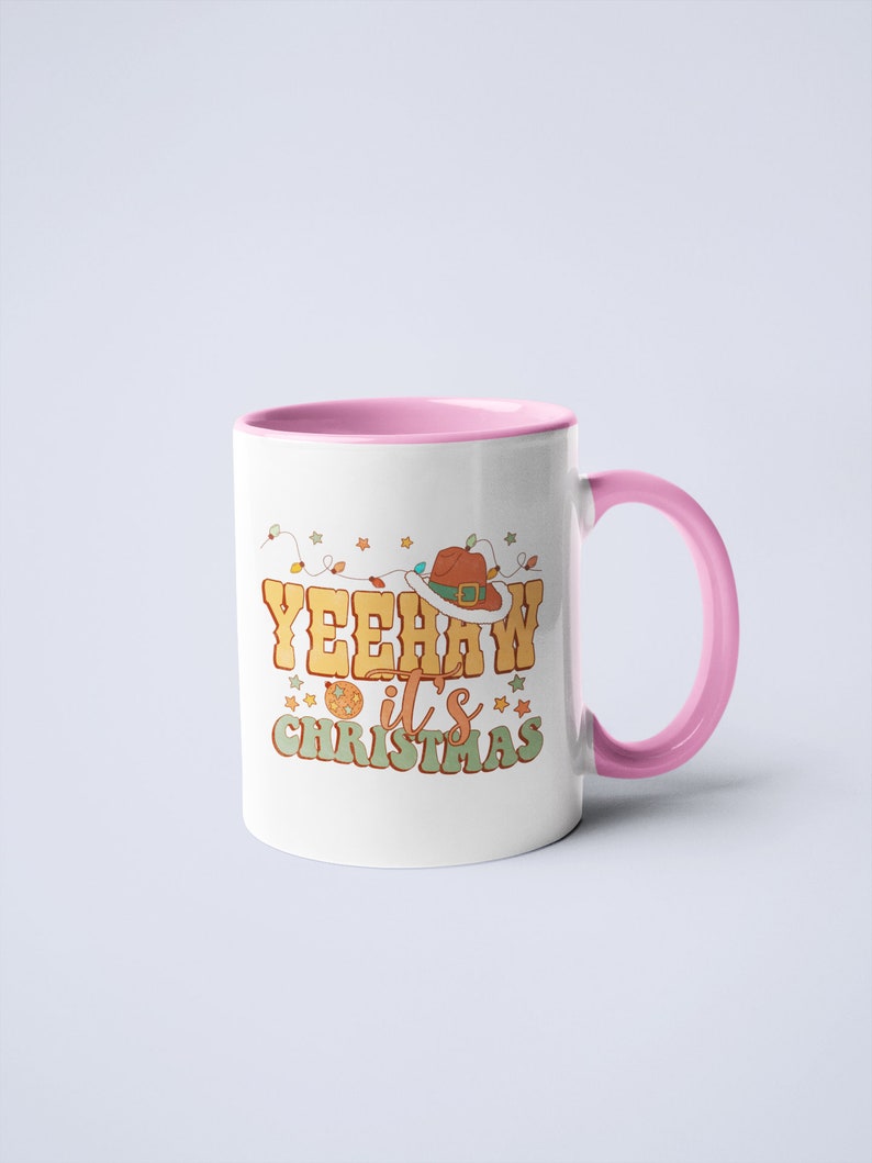 May include: A white ceramic mug with a pink interior and handle. The front features a graphic with the text YEEHAW it's CHRISTMAS in yellow and green letters, accompanied by a brown cowboy hat, a disco ball, and colorful string lights.