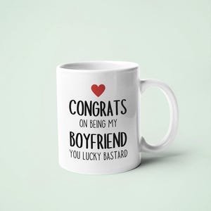 A white ceramic mug with black text that reads "CONGRATULATIONS ON BEING MY BOYFRIEND YOU LUCKY BASTARD" and a red heart symbol.