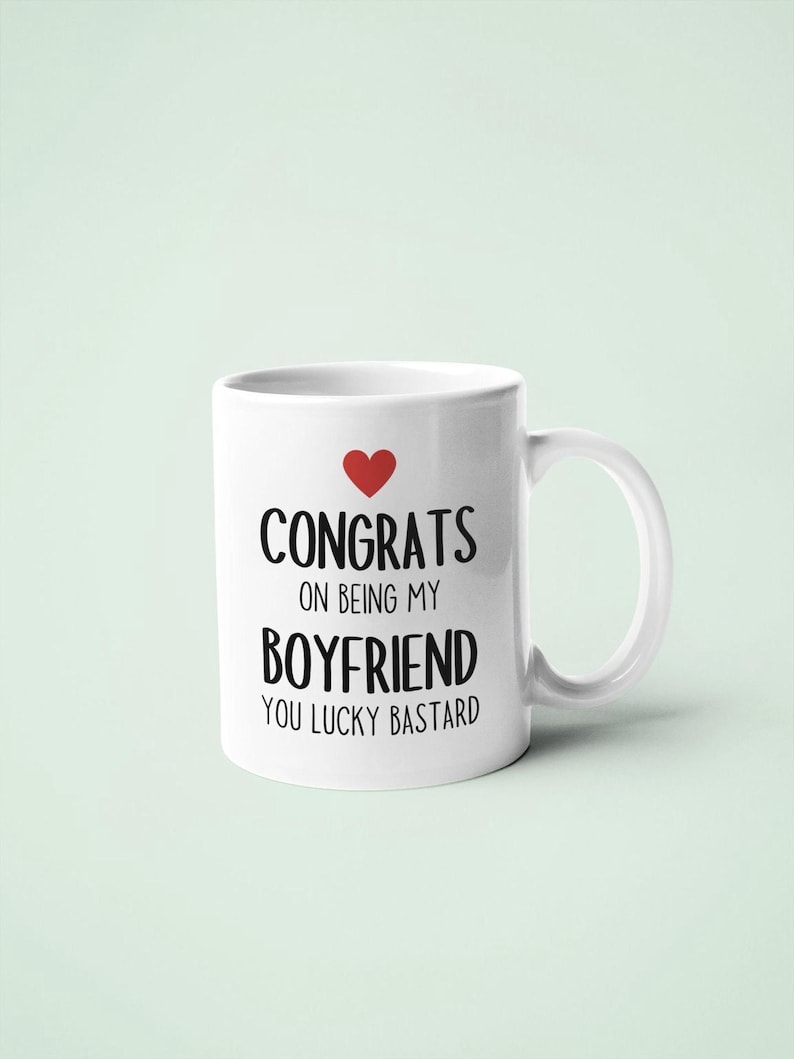 A white ceramic mug with black text that reads "CONGRATULATIONS ON BEING MY BOYFRIEND YOU LUCKY BASTARD" and a red heart symbol.