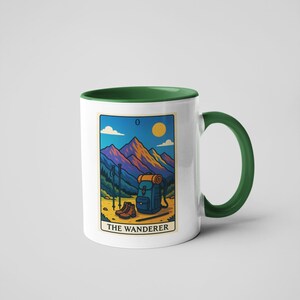 May include: White ceramic mug with a green handle and interior. The mug features a colorful illustration of a mountain scene with hiking gear and the text "THE WANDERER". The design includes a backpack, boots, and hiking poles.