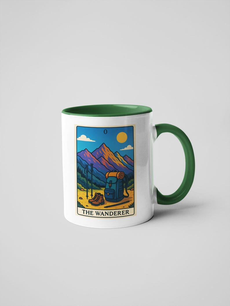 May include: White ceramic mug with a green handle and interior. The mug features a colorful illustration of a mountain scene with hiking gear and the text "THE WANDERER". The design includes a backpack, boots, and hiking poles.