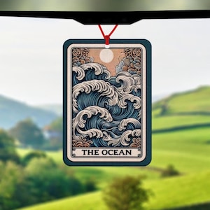 May include: Rectangular air freshener with a blue border and a red string for hanging. The design features a detailed illustration of ocean waves, a sun, and the text "THE OCEAN". The background shows a green landscape.
