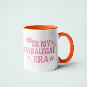 Op de afbeelding: Witte keramische mok met een oranje binnenkant en handvat. De mok heeft de tekst "IN MY PARALEGAL ERA" in roze, met bloemen- en sterrenafbeeldingen. De mok staat tegen een lichtblauwe achtergrond.