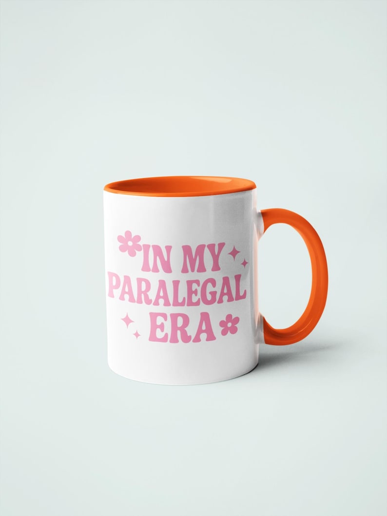 Op de afbeelding: Witte keramische mok met een oranje binnenkant en handvat. De mok heeft de tekst "IN MY PARALEGAL ERA" in roze, met bloemen- en sterrenafbeeldingen. De mok staat tegen een lichtblauwe achtergrond.