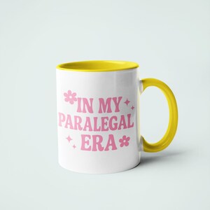 Op de afbeelding: Witte keramische mok met een gele binnenkant, oor en rand. De mok heeft de tekst "IN MY PARALEGAL ERA" in roze, met bloem- en sterrenafbeeldingen. Ontworpen voor dranken.