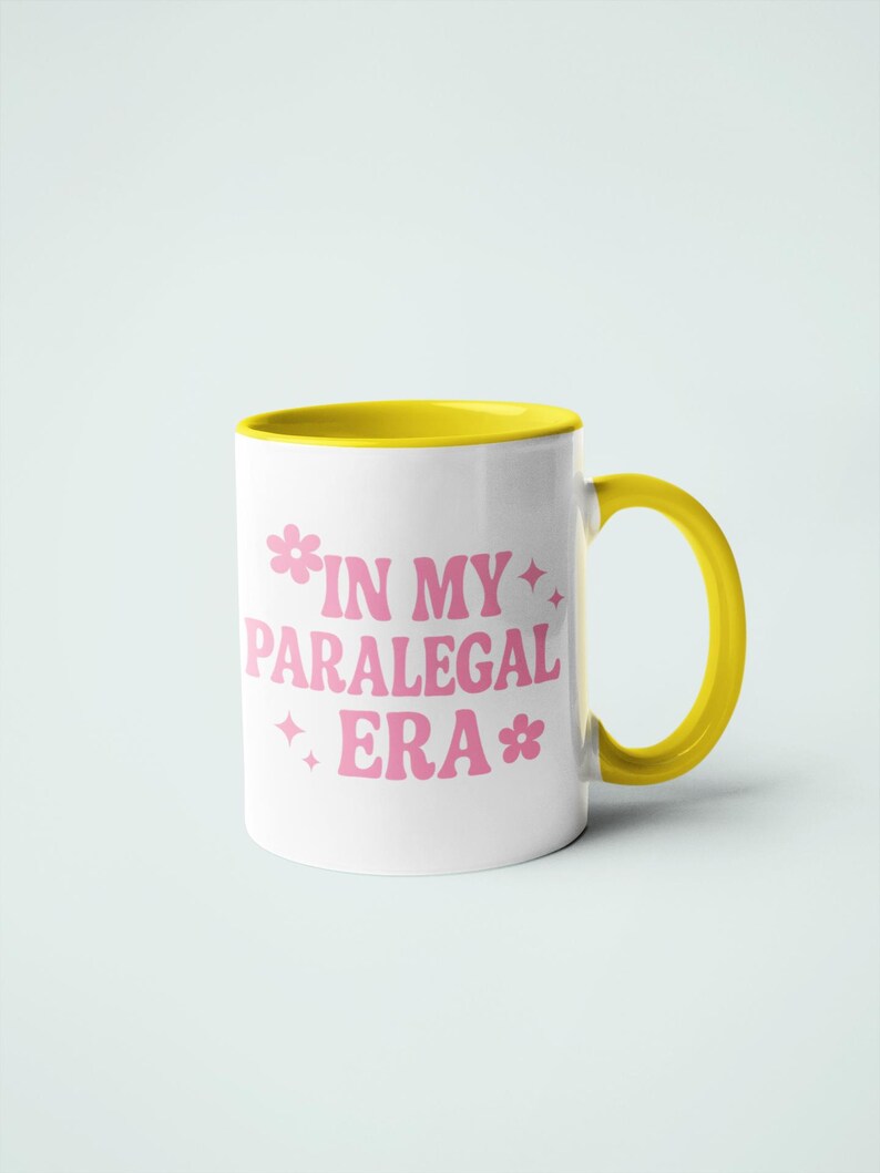 Op de afbeelding: Witte keramische mok met een gele binnenkant, oor en rand. De mok heeft de tekst "IN MY PARALEGAL ERA" in roze, met bloem- en sterrenafbeeldingen. Ontworpen voor dranken.