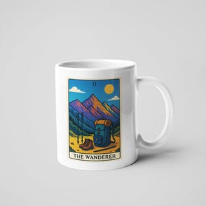 May include: White ceramic mug featuring a colorful illustration of a mountain scene with hiking gear. The image includes a backpack, hiking boots, and trekking poles. The text "THE WANDERER" is displayed below the image.