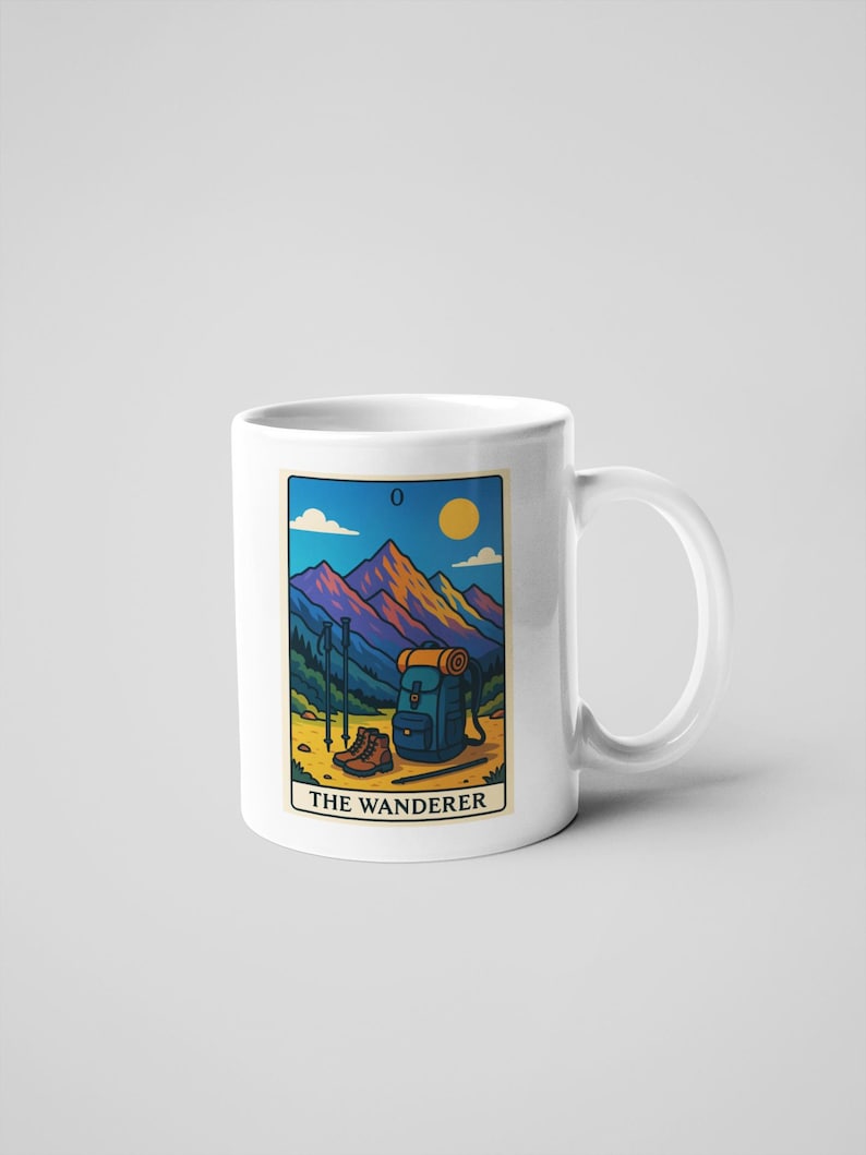 May include: White ceramic mug featuring a colorful illustration of a mountain scene with hiking gear. The image includes a backpack, hiking boots, and trekking poles. The text "THE WANDERER" is displayed below the image.