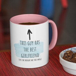 Pode incluir: Caneca de cer&acirc;mica branca com borda e al&ccedil;a rosa. A caneca tem um design de aquarela azul e texto preto que diz "Este cara tem a melhor namorada (Sim, ela me comprou esta caneca)".