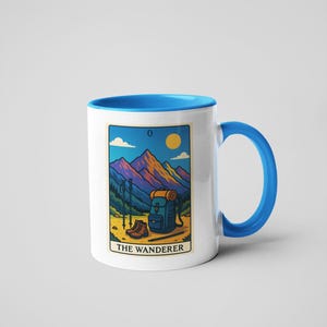 May include: White ceramic mug with a blue interior and handle. The mug features a colorful illustration of a mountain scene with hiking gear and the text "THE WANDERER". The design includes a backpack, hiking boots, and trekking poles.