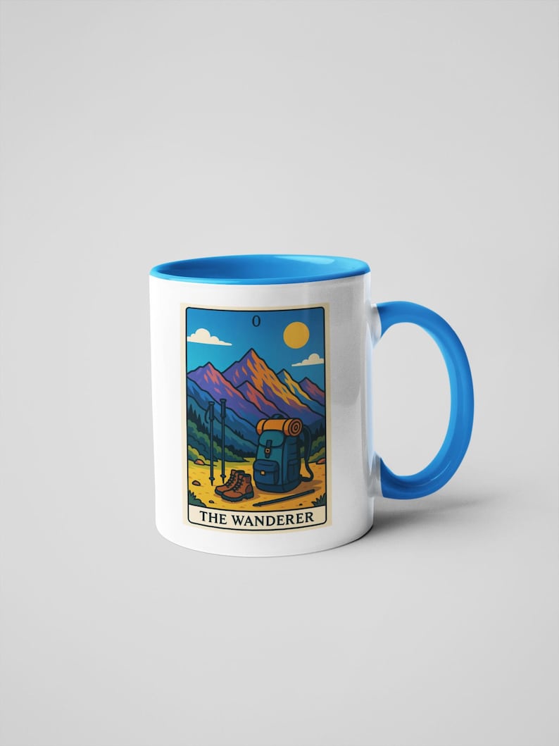 May include: White ceramic mug with a blue interior and handle. The mug features a colorful illustration of a mountain scene with hiking gear and the text "THE WANDERER". The design includes a backpack, hiking boots, and trekking poles.