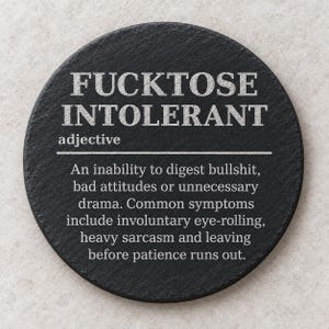 May include: A round black slate coaster featuring white engraved text that reads FUCKTOSE INTOLERANT, defined as an adjective describing an inability to digest bullshit, bad attitudes, or drama, with symptoms like eye-rolling and heavy sarcasm.