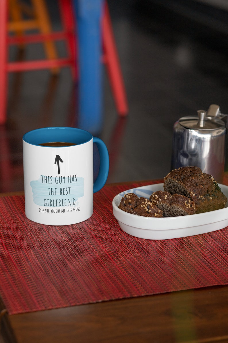 Pode incluir: Caneca de caf&eacute; branca com borda e al&ccedil;a azuis. A caneca tem um design de aquarela azul e branco com o texto "Este cara tem a melhor namorada (Sim, ela me comprou esta caneca)" e uma seta apontando para cima.
