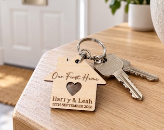 May include: A light-colored wooden keychain shaped like a house with a heart cutout. The text "Our First Home" is engraved at the top, with "Harry & Leah" and "12TH SEPTEMBER 2026" below. A silver metal keyring holds two standard metal keys.