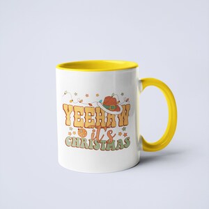 May include: A white ceramic mug with a bright yellow interior and handle. The front features a graphic with the text YEEHAW it's CHRISTMAS in orange and green letters, accompanied by a brown cowboy hat, string lights, and small star icons.