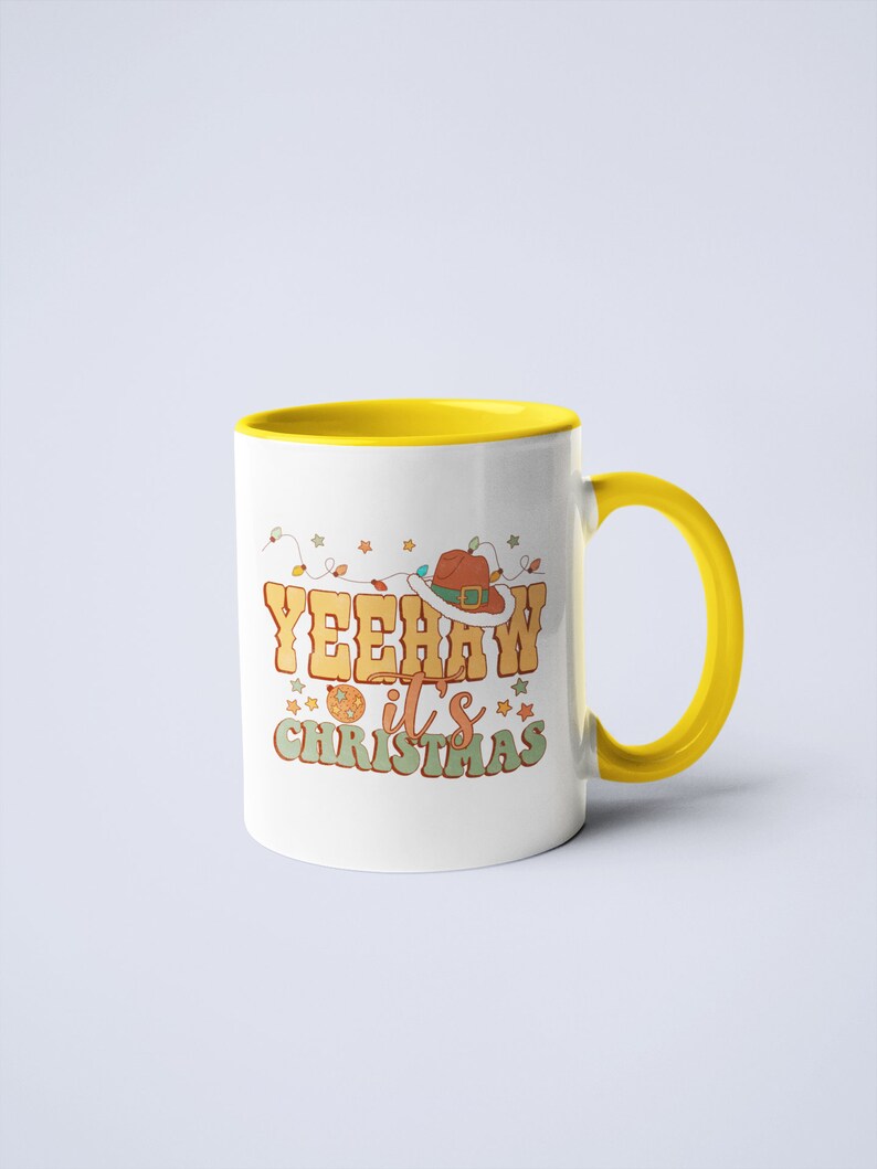May include: A white ceramic mug with a bright yellow interior and handle. The front features a graphic with the text YEEHAW it's CHRISTMAS in orange and green letters, accompanied by a brown cowboy hat, string lights, and small star icons.