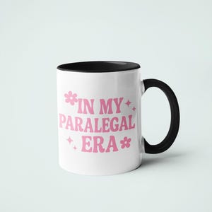 Op de afbeelding: Witte keramische mok met een zwarte binnenkant en handvat. De mok heeft de tekst "IN MY PARALEGAL ERA" in roze, met bloem- en steraccenten. Ontworpen voor dranken.