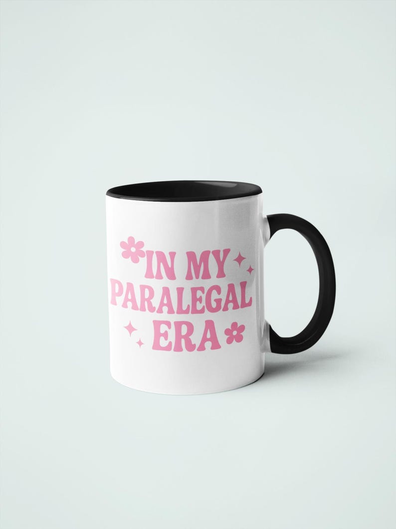 Op de afbeelding: Witte keramische mok met een zwarte binnenkant en handvat. De mok heeft de tekst "IN MY PARALEGAL ERA" in roze, met bloem- en steraccenten. Ontworpen voor dranken.