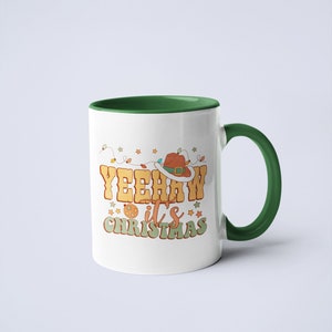 May include: A white ceramic mug with a dark green interior and handle. The front features a graphic with the text YEEHAW it's CHRISTMAS in retro-style lettering, accompanied by a brown cowboy hat, string lights, and small star icons.