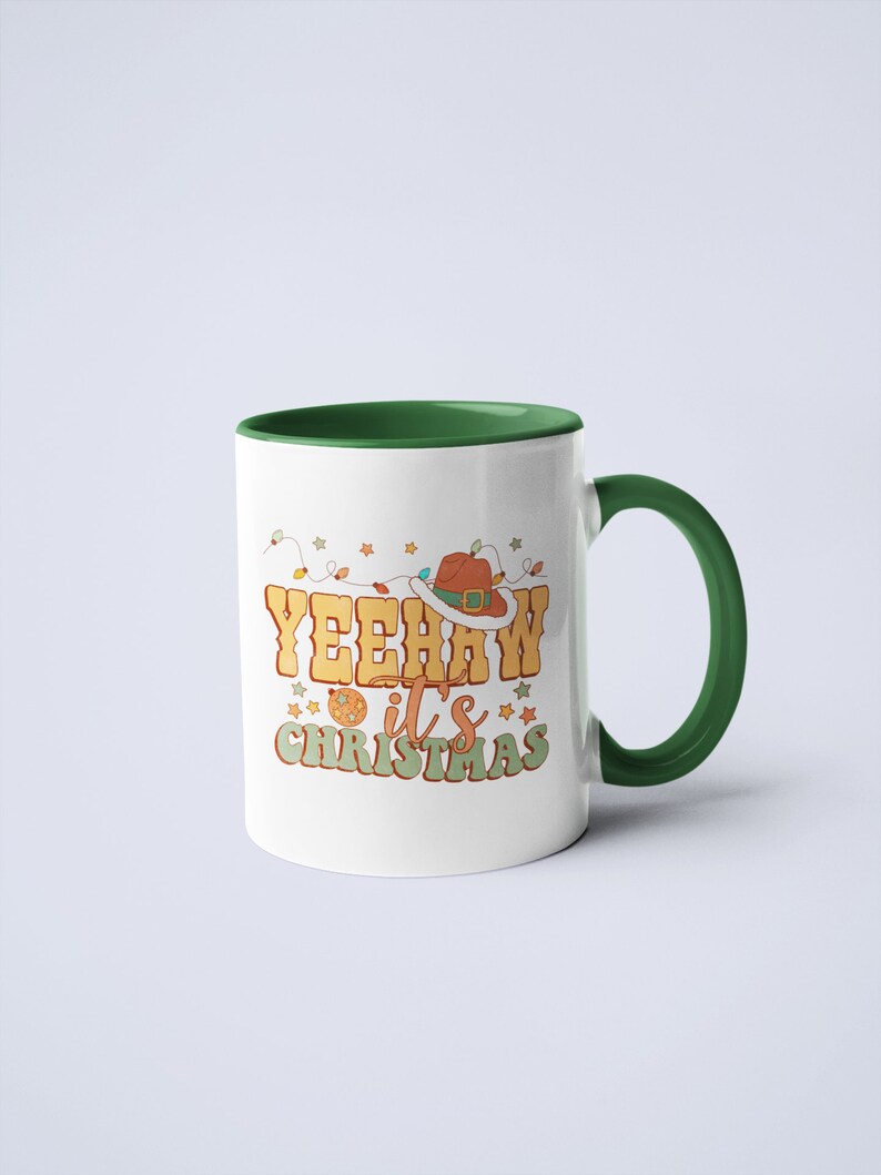May include: A white ceramic mug with a dark green interior and handle. The front features a graphic with the text YEEHAW it's CHRISTMAS in retro-style lettering, accompanied by a brown cowboy hat, string lights, and small star icons.
